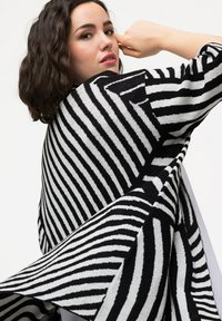 Stickad omslag med kraftiga svartvita ränder, som visar en flödande design, texturerad tyg och breda ärmar för en lagerlook.