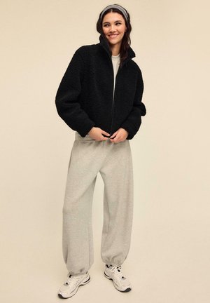 Crna flis jakna s visokim ovratnikom, sive trenirka hlače i bijele sportske tenisice. Glatke teksture, opušteni kroj i jednostavni dizajnerski elementi.