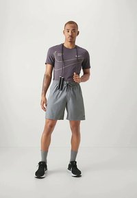 Urheilullinen mies harmaassa Nike-t-paidassa ja shortseissa, mustissa lenkkareissa, harmaissa sukissa, pidellen mustaa hyppynarua yksinkertaista valkoista taustaa vasten.