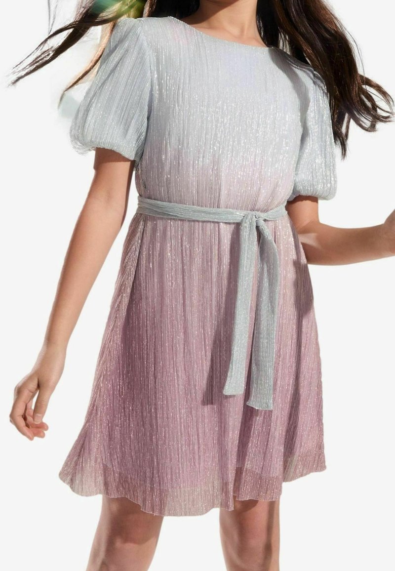 Fille portant une robe courte scintillante rose et argent avec une ceinture argentée nouée à la taille, debout sur un fond blanc.