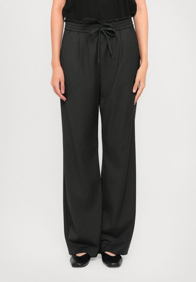 Personne portant un pantalon noir ample à cordon de serrage avec des plis, associé à des chaussures plates noires sur un fond clair.