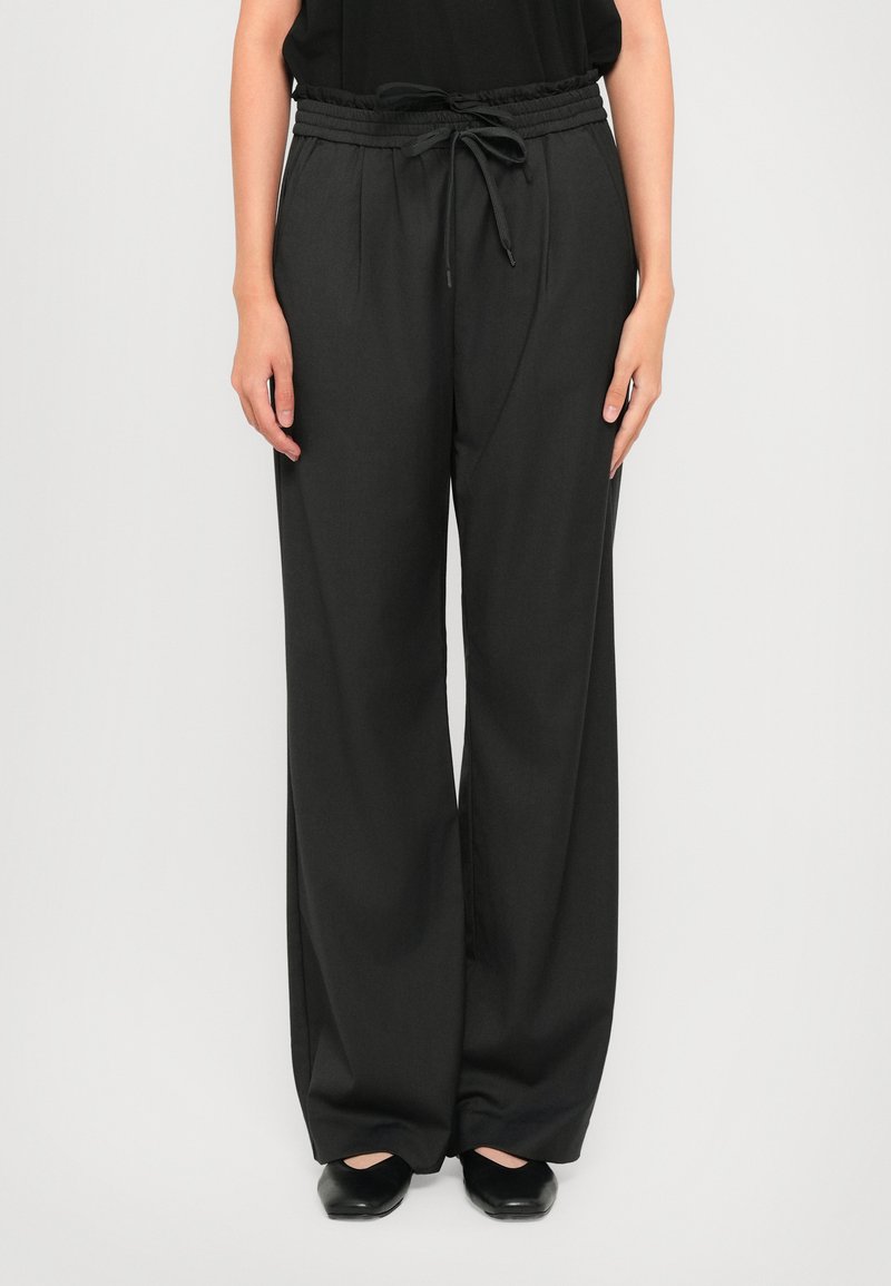 Person wearing loose black drawstring pants with pleats, paired with black flat shoes on a plain light background.