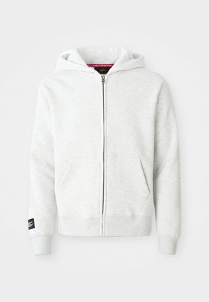 Svijetlosivi zip-up dukserica od mekog materijala, s kapuljačom, prednjim džepovima i crnom oznakom na lijevom rukavu.