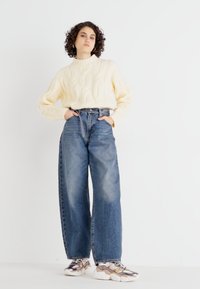 Cream cable-knit sweater, high-waisted blue wide-leg jeans, and chunky sneakers with metallic accents. Plain background.