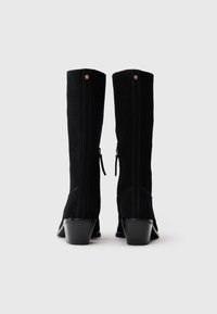 Μαύρες σουέτ μπότες μέχρι το γόνατο με χοντρό τακούνι, που διαθέτουν φερμουάρ και διακοσμητική ραφή στο πίσω μέρος. Χρυσή λεπτομέρεια στην κορυφή.