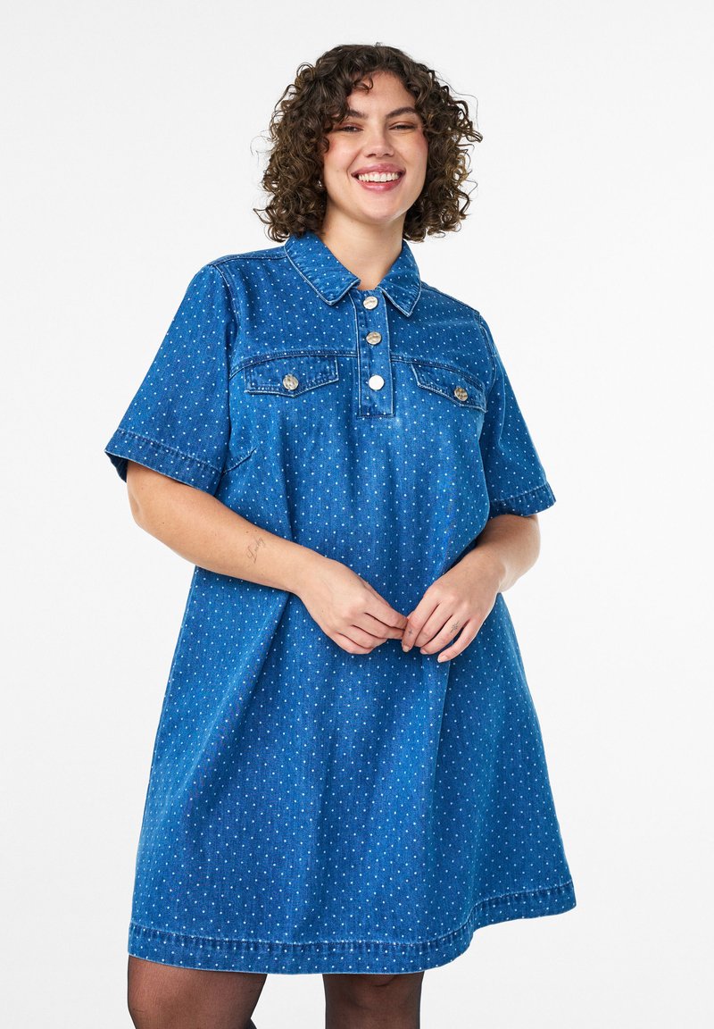 Robe en denim bleue avec manches courtes, col, deux poches poitrine et pois blancs. Elle présente des boutons ronds et une silhouette évasée.