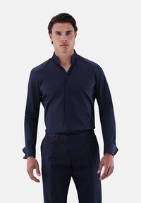 Dunkelblauses Hemd mit Stehkragen, strukturiertem Stoff und langen Ärmeln. Getragen mit einer dunklen Hose, die einen Knopfdetails an der Taille aufweist.