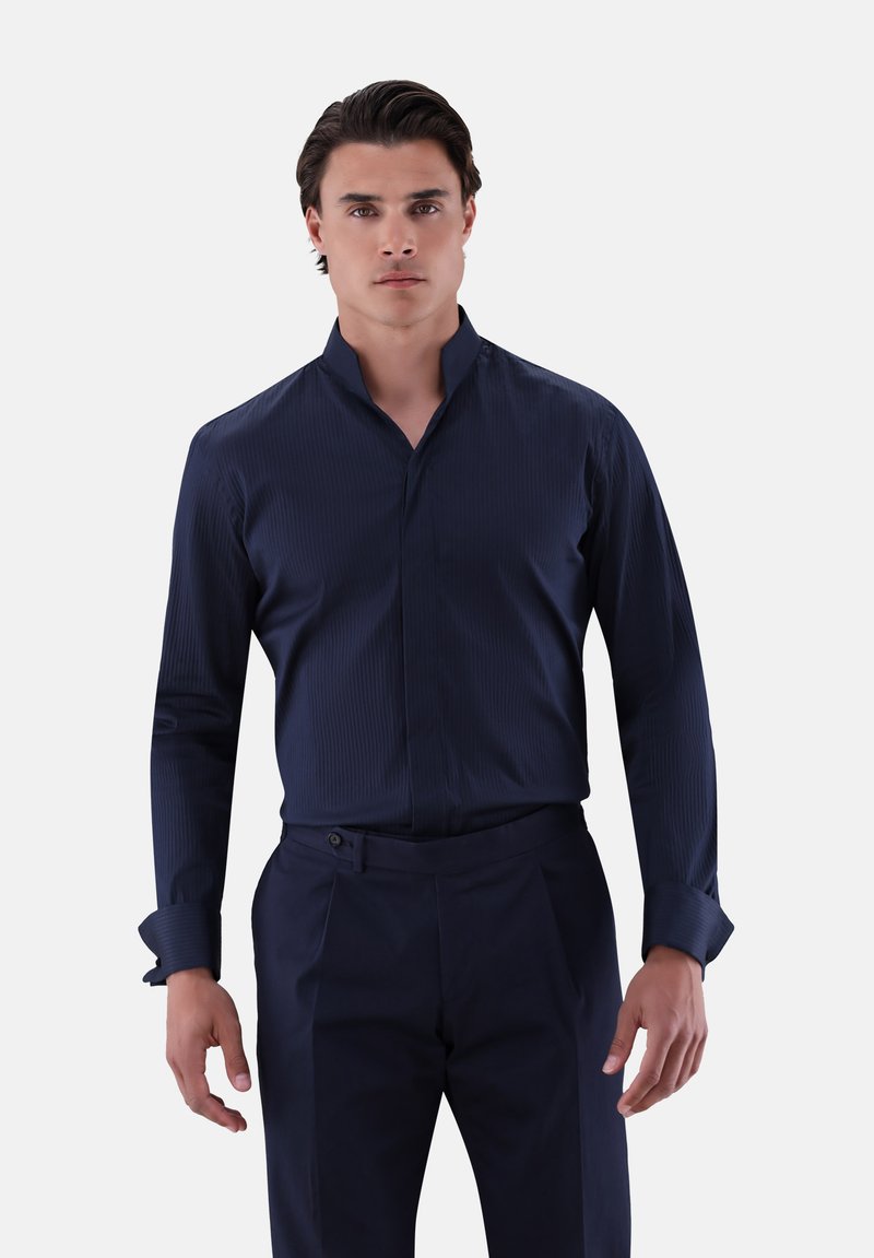 Dunkelblauses Hemd mit Stehkragen, strukturiertem Stoff und langen Ärmeln. Getragen mit einer dunklen Hose, die einen Knopfdetails an der Taille aufweist.