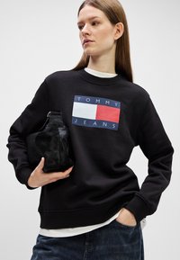 Mustat collegepaidat, jossa on suorakulmainen logo punaisista, valkoisista ja sinisistä osista. Malli pitää käsissään teksturoitua mustaa käsilaukkua. Rentoa tyyliä.