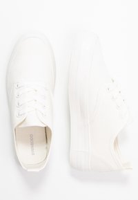Sneakers bianchi in tela con punta arrotondata, lacci piatti e suola in gomma testurizzata con lievi strisce verticali sul tallone.