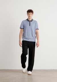 Camisa polo azul clara com gola escura, textura canelada e detalhe de logótipo, combinada com calças pretas e sapatilhas brancas. Fundo neutro.