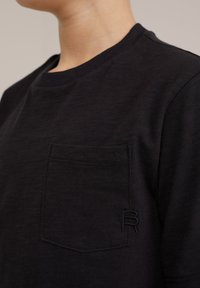 Čierne tričko s krátkym rukávom vyrobené z texturovanej látky. Obsahuje náprsné vrecko a vyšívané logo na boku. Jednoduchý okrúhly výstrih.