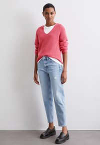 Roze gebreide trui met een diepe V-hals, laag over een witte T-shirt; gecombineerd met lichtblauwe versleten jeans en zwarte instappers.