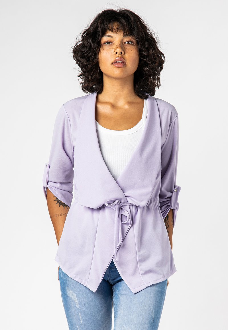 Lavendel wikkeltop met driekwartmouwen, een strik in de taille en zachte, gladde stof; gecombineerd met een witte blouse en lichtblauwe jeans.