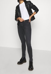 Černá kožená motorkářská bunda přes bílý tričko, spárovaná s tmavě šedými úzkými džíny a černými šněrovacími botami. Jednoduchý, moderní design.