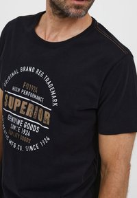 Melna īsām piedurknēm t-krekls ar apaļu kakla izgriezumu, ar zelta un baltā tekstā izdrukātu dizainu, kas izceļ "SUPERIOR" un dibināšanas gadu.