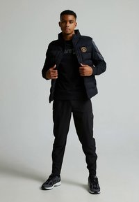 Svart boblejakke med grå detaljer, glidelås foran, logo på brystet, kombinert med svart t-skjorte og svarte bukser, fullført med svarte joggesko.