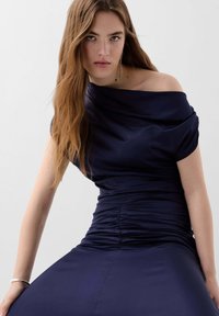 Laivaston sininen off-shoulder-mekko, jossa on laskostettua kangasta, rypytystä vyötäröllä ja sileä, vartalonmyötäinen siluetti. Teksturoitu, pehmeä materiaali.