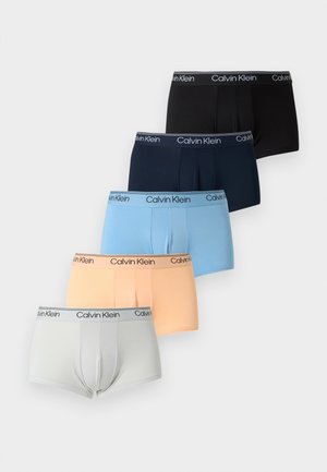 Νεαρός άντρας, όρθιος ξυπόλητος με μαύρα boxer Calvin Klein μπροστά από ένα σκούρο πράσινο δερμάτινο καναπέ σε μπεζ χαλί.