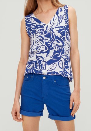 Femme portant un haut sans manches floral bleu et blanc associé à un short en denim bleu retroussé, devant un fond uni.