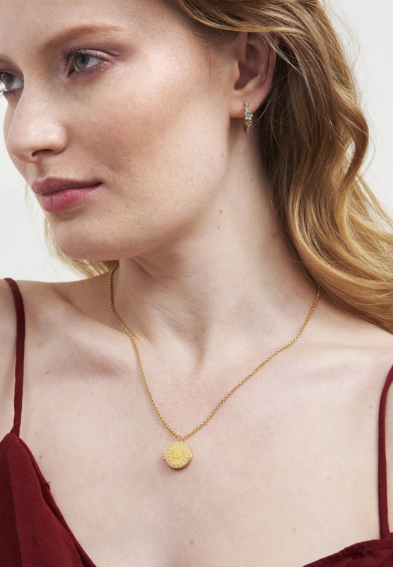 Gouden halsketting met een rond patroonhanger aan een fijne ketting, gedragen op een blote schouder, aangevuld met een oorbel met turquoise accenten.