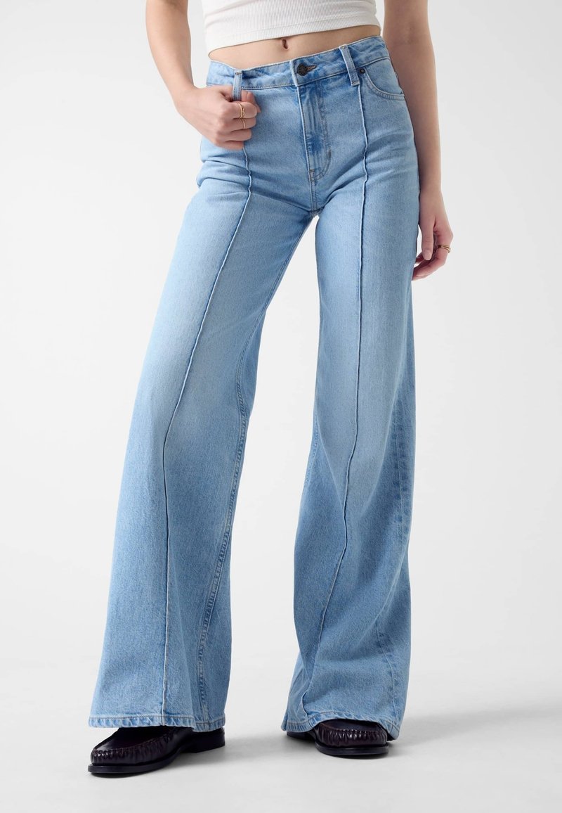 Jeans évasés en denim bleu clair à taille haute avec surpiqûres contrastantes, fermeture à boutons et jambes larges. La surface a une texture lisse.