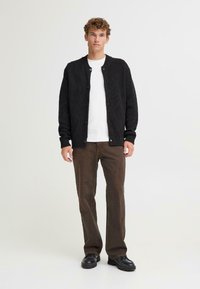 Cardigan nero a coste con scollatura rotonda e chiusura a bottoni, indossato sopra una maglietta bianca, abbinato a pantaloni marroni e scarpe nere.