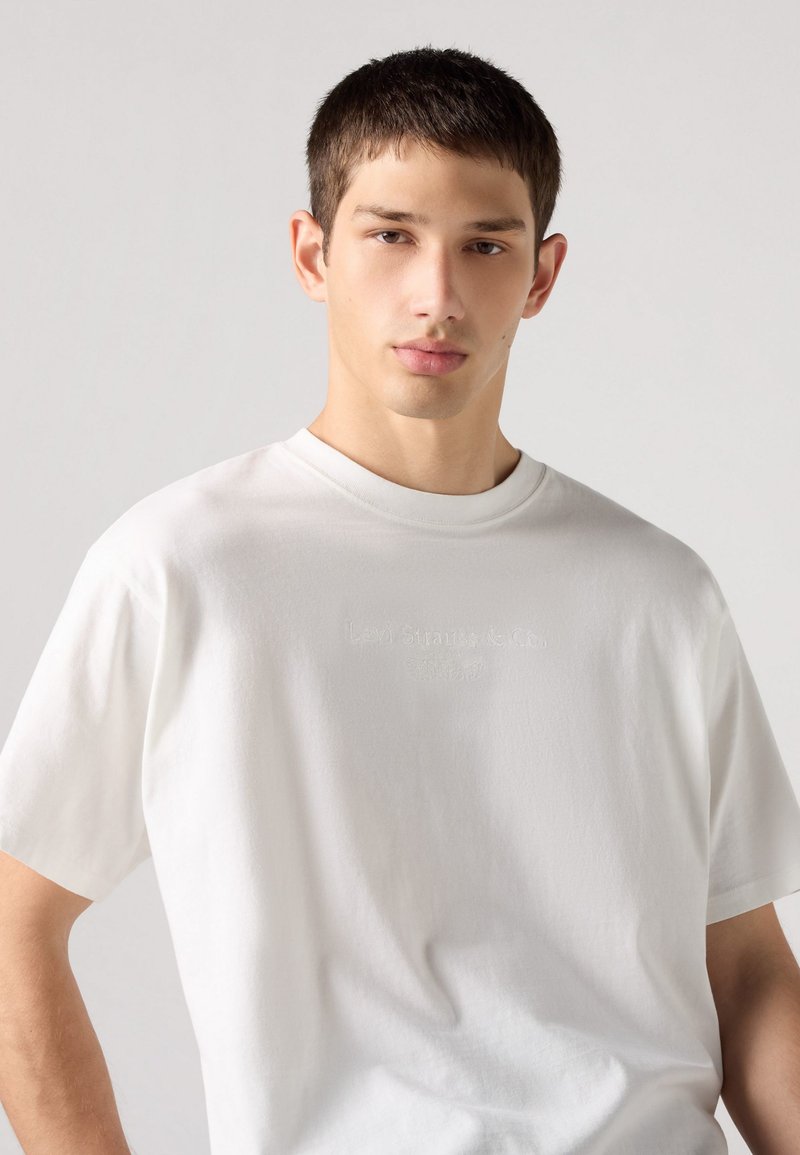 Weißes Baumwoll-T-Shirt mit kurzen Ärmeln, das ein erhabenes Logo in der Mitte aufweist. Der Stoff hat eine glatte Textur und einen lockeren Schnitt.