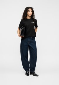 Μαύρο βαμβακερό t-shirt με λογότυπο, φαρδιά μπλε τζιν, και μαύρα δερμάτινα μποτάκια μέχρι τον αστράγαλο. Το μοντέλο κρατάει μια μαύρη τσάντα χειρός.