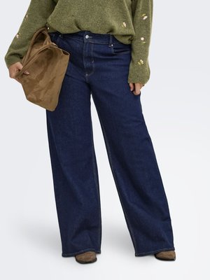 Person iført mørkeblå vidde jeans, olivengrøn sweater med lille blomsterbroderi, holder en stor brun ruskindstaske.