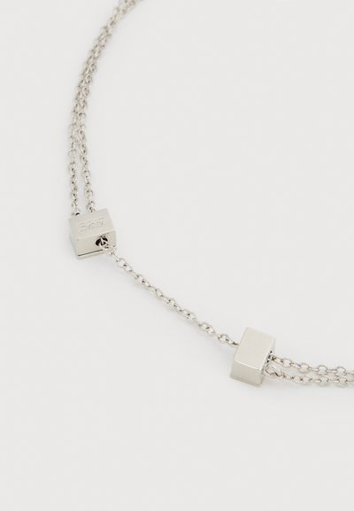 Διπλό ασημένιο κολιέ με δύο μικρές ασημένιες κύβους, μία από τις οποίες είναι χαραγμένη με τη λέξη "VERSACE", σε λευκό φόντο.