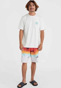 Camiseta blanca de algodón con logo circular azul, combinada con bañador multicolor que presenta un patrón degradado de rojo, naranja, amarillo y azul.