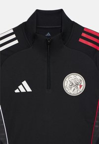 Svart träningsjacka med zip-kra med vita och röda ränder på axlarna samt en Ajax-logotyp i en cylindrisk lapp. Slät textur.