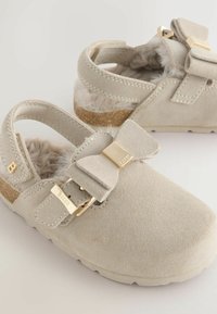 Des chaussures gris clair avec un détail en noeud et des accents dorés, présentant une doublure douce et duveteuse, mises en valeur sur un fond clair.