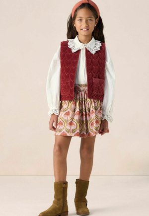 Jeune fille portant un gilet matelassé rouge, une blouse blanche avec un col en dentelle, une jupe à fleurs, des bottes marron et un bandeau rouge, se tenant devant un fond uni.