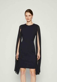 Μαύρο φόρεμα με διαφάνειες μανίκια σαν κάπα, εφαρμοστή σιλουέτα, στρογγυλή λαιμόκοψη και μήκος ως το γόνατο. Λείο ύφασμα με δομημένο σχέδιο.