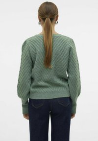 Maglione verde lavorato a maglia con motivo diagonale strutturato, maniche ampie e polsini a costine, abbinato a jeans di denim scuro.