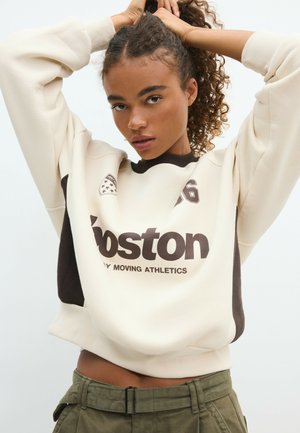 Jeune femme aux cheveux bouclés portant un sweat-shirt Boston blanc et noir et un pantalon vert taille haute, levant ses cheveux avec les deux mains.