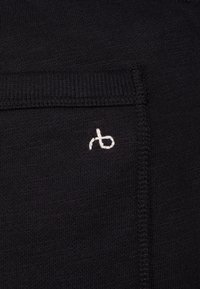 Schwarzer Stoff mit feiner Textur, der ein kleines, weißes, gesticktes Logo zeigt. Das Design umfasst eine sichtbare Naht entlang der oberen Kante.