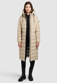 Beige lange pufferjas met een capuchon, voorzien van doorgestikte vulling, een ritssluiting aan de voorkant en zijzakken. Draagt over een zwarte broek en laarzen.