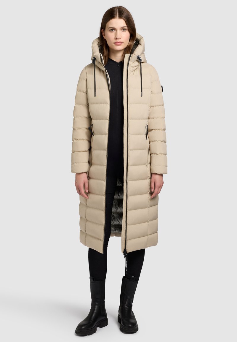 Beige lange pufferjas met een capuchon, voorzien van doorgestikte vulling, een ritssluiting aan de voorkant en zijzakken. Draagt over een zwarte broek en laarzen.