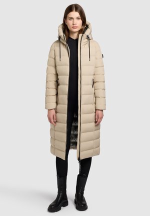Beige pufferjas met capuchon, met een gestikt ontwerp, ritssluiting en zijzakken. Draag het over een zwarte outfit en gecombineerd met zwarte laarzen.
