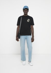 Camiseta negra de manga corta con un gráfico en el frontal, jeans de mezclilla sueltos de color azul claro, zapatillas blancas y una gorra azul, de pie contra un fondo blanco.