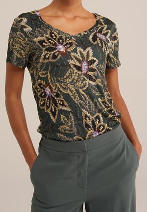 Kobieta w ciemnej bluzce z krótkim rękawem w kwiatowy wzór, wpuszczonej do wysokowasu spodni w stonowanym zielonym kolorze, z rękami w kieszeniach.