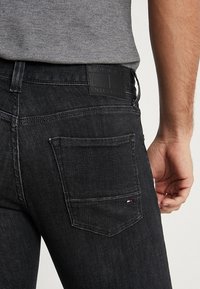 Svarta denimjeans har en texturerad yta, med en läderpatch vid bakens midja och en liten logodetalj på bakfickan.