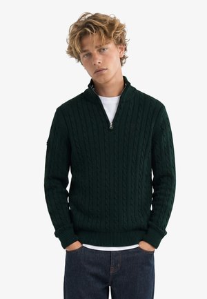 Jonge man met blond krullend haar die een donkergroene kabelgebreide trui met halve rits, een wit T-shirt en donkerblauwe jeans draagt, handen in de zakken.