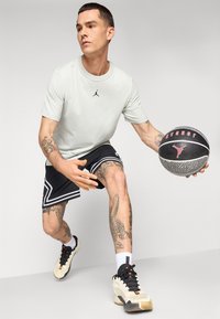 Mænds lys grå T-shirt med sort Jordan-logo, sorte shorts med hvide accenter og mønstre, holder en sort basketball med røde detaljer.
