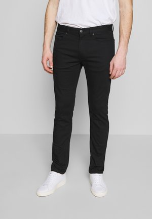 Homme portant un jean noir slim, des baskets blanches et un t-shirt blanc uni, debout devant un fond gris.