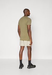 Olive green short-sleeve t-shirt with a round neckline, paired with light beige shorts. The model wears black socks and athletic shoes.
