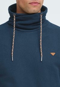 Uomo che indossa un felpa blu navy con colletto alto e cordini multicolori, dotata di un piccolo logo triangolare marrone sul petto.